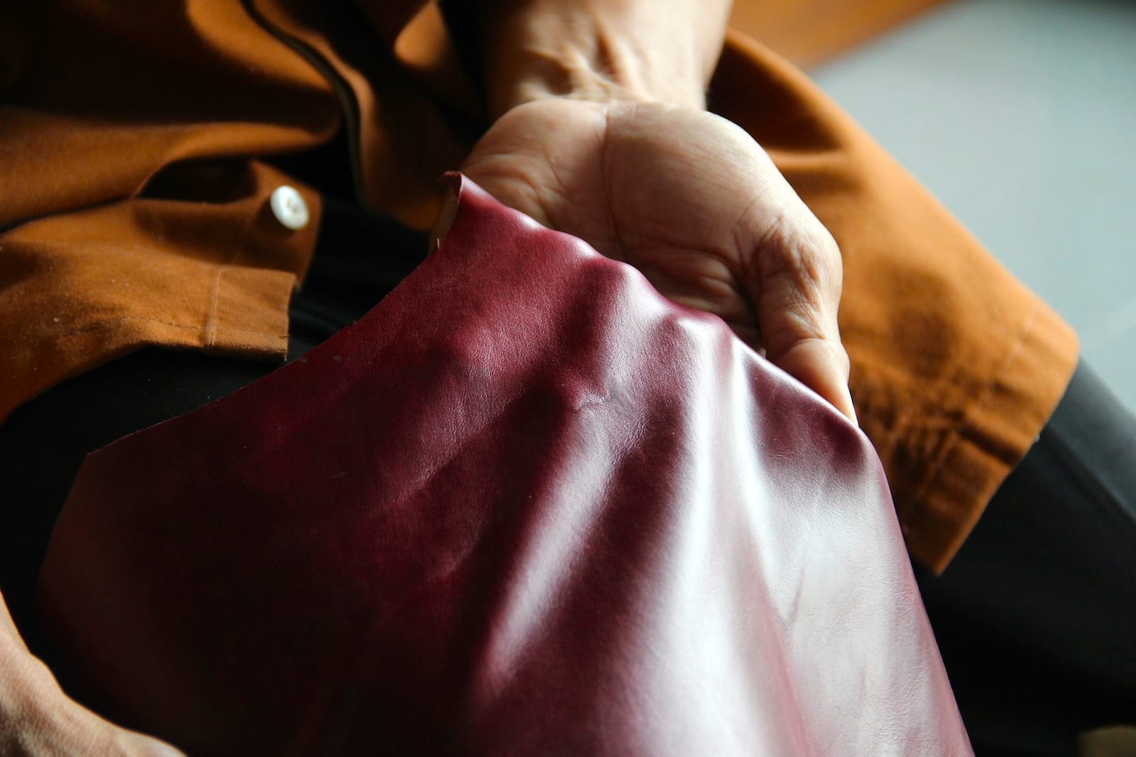Tangan menggenggam selembar kulit burgundy yang halus, lembarannya terlipat lembut dengan kilap tipis